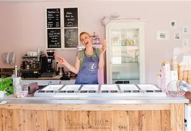 A friendly ice cream shop employee stands behind the ice cream cart and holds an ice cream in her hand. The background is light pink, featuring a selection of ice cream flavors and various utensils.
