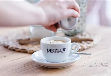 A cup of coffee with cream on a small plate. In the background, sugar is being added.