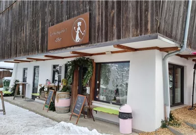 A cozy café with a rustic wooden façade and large windows. The entrance area is inviting with plants and outdoor tables.