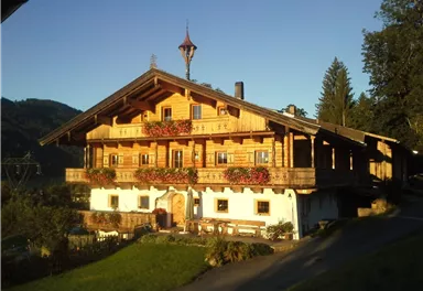 A traditional wooden house in Alpine style, surrounded by green meadows and mountains. The balconies are adorned with colorful flowers, and the sun is shining on the building.