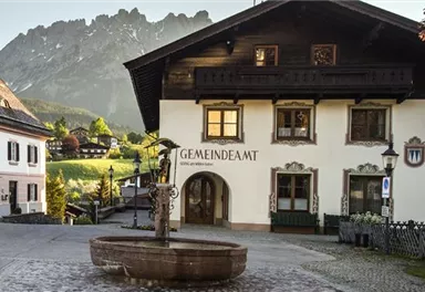A picturesque village with traditional houses and a beautiful fountain. In the background, impressive mountains and a green landscape can be seen.