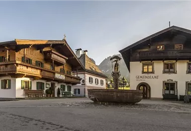 A picturesque village square with traditional wooden buildings. In the center, there is a beautiful fountain, surrounded by a tranquil landscape.
