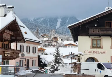 A picturesque winter landscape with snow-covered roofs and mountains in the background. In the foreground, traditional buildings and a vehicle can be seen.