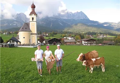 A picturesque rural scene with three people in traditional clothing standing in front of a church. In the foreground, two cows can be seen on a green meadow, surrounded by mountains.