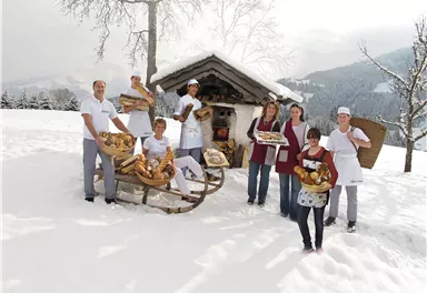 A group of people stands in the snow in front of a small cabin. They are holding fresh bread and pastries in their hands.
