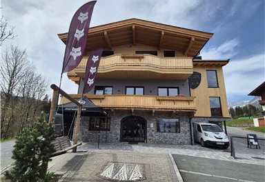 Ein modernes Gebäude mit Holzverkleidung und großen Fenstern. Davor stehen Fahnen und ein Parkplatz ist vorhanden.