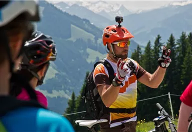 Ein Radtrainer erklärt eine Technik an einem sonnigen Tag. Im Hintergrund sind Berge und Bäume zu sehen.