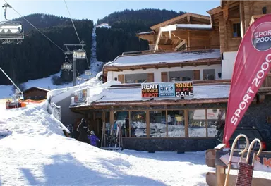 A snowy area with a mountain restaurant and ski rental. In the background, ski lifts and snow-covered mountains can be seen.