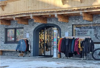 Ein Sportgeschäft mit einem einladenden Eingang. Vor dem Laden hängen verschiedene Kleidungsstücke auf Ständern.