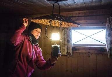 Eine Person steht in einem Holzraum und hält eine Lampe über einen Korb. Es ist dunkel, aber das Licht der Lampe beleuchtet den Raum.