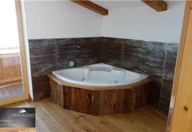 Ein modernes Badezimmer mit einer luxuriösen Eckbadewanne aus Holz. Die Wände sind in natürlichen Farbtönen gehalten, und große Fenster sorgen für viel Tageslicht.