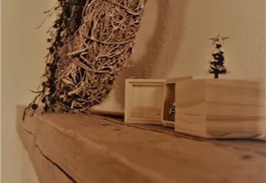Ein dekorativer Holzregal mit einem Kranz aus natürlichen Materialien und einer kleinen Holzbox. Daneben steht ein kleiner Weihnachtsbaum.