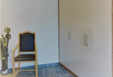 Ein minimalistischer Raum mit einem Holzstuhl und einem großen, weißen Schrank. Die Wände sind hell, und der Boden ist aus grauem Stein.