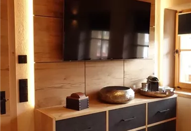 Ein modernes Wohnzimmer mit einem Fernseher an der Wand und einer eleganten Holzkommode. Die Kommode hat schwarze Schubladen und ist mit Dekorationsobjekten geschmückt.