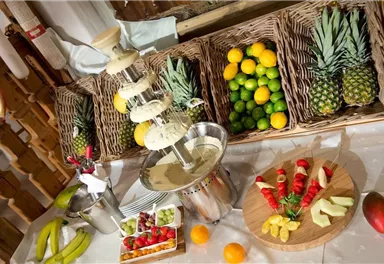 A chocolate fountain with fresh fruits and a beautiful presentation. In the background, there are fruit baskets with pineapples, limes, and oranges.