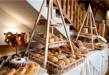 A buffet with freshly baked bread and pastries. On wooden shelves, various types of bread and a pretzel catching station can be seen.
