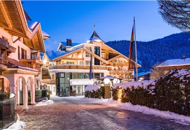 A picturesque Alpine hotel in winter at dusk. The snow blankets the path and the surrounding trees, while the lights of the building glow warmly.