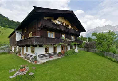 A traditional wooden house with a balcony, surrounded by a green meadow and mountains in the background. The idyllic setting invites relaxation.