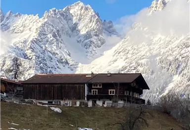 A traditional wooden house is picturesque in front of an impressive mountain landscape. The snow-capped peaks form a beautiful contrast to the green slope.