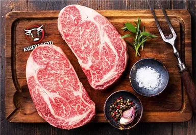 Two large pieces of high-quality beef on a wooden board, surrounded by spices and fresh herbs. Next to it is a spatula knife and salt in a small bowl.