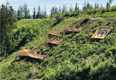 Eine grüne Hügellandschaft mit terrassierten Holzstegen. Im Hintergrund ist ein Kunstwerk an der Wand sichtbar.