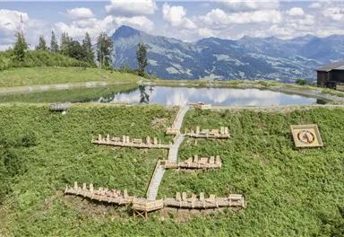 Eine malerische Landschaft mit einer Holzliegefläche inmitten von grünen Wiesen und einem ruhigen Teich. Im Hintergrund sind majestätische Berge und ein blauer Himmel zu sehen.