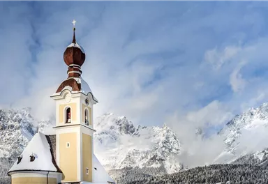 A picturesque church with a distinctive onion dome spire, surrounded by snow-covered mountains. The sky is clear and the landscape appears peaceful and wintry.