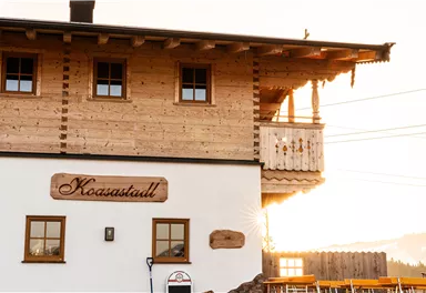 Ein holzverkleidetes Gebäude mit dem Namen "Koaastall". Im Hintergrund ist die Abendsonne zu sehen, die für eine warme Atmosphäre sorgt.