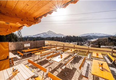 Eine sonnige Terrasse mit Holztischen und Bänken bietet einen Blick auf die Berge. Im Hintergrund ist eine klare, blaue Himmelslandschaft zu sehen.