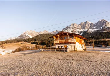 Ein schönes Holzhaus steht in einer winterlichen Landschaft mit schneebedeckten Bergen im Hintergrund. Die Sonne scheint und schafft eine friedliche Atmosphäre.