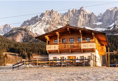 Ein traditionelles Holzhaus in den Bergen, umgeben von schneebedeckten Gipfeln. Die Landschaft ist friedlich und idyllisch.