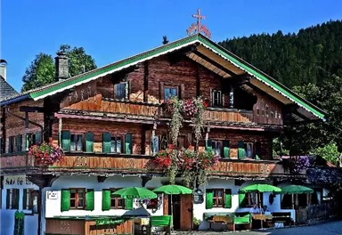 Ein traditionelles alpenländisches Haus aus Holz mit blühenden Pflanzen und grünen Fenstern. Vor dem Gebäude stehen Sitzgelegenheiten mit Sonnenschirmen.