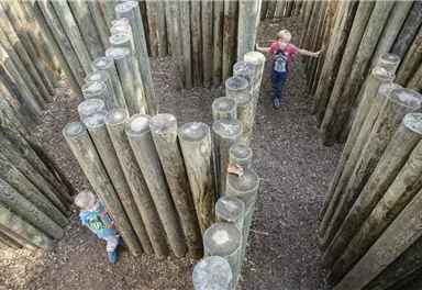 Ein Spielplatzlabyrinth aus Holzstämmen mit zwei Kindern, die darin spielen. Die Umgebung ist naturbelassen und einladend.