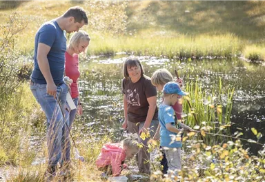 Eine Familie steht am Ufer eines Teiches und angelt. Kinder spielen im Wasser und Erwachsene beobachten sie.