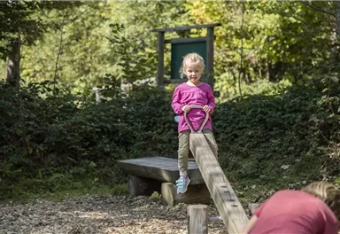 Ein Mädchen steht auf einer Wippe im Spielplatz. Es ist ein sonniger Tag umgeben von Bäumen.