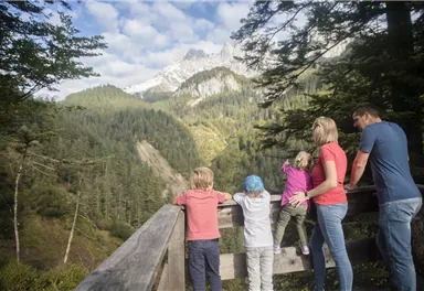 Eine Familie steht an einer Aussichtsplattform und blickt auf eine beeindruckende Berglandschaft. Im Hintergrund sind grüne Wälder und schneebedeckte Gipfel zu sehen.