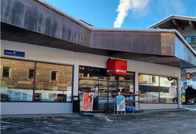 Ein geöffneter Laden mit großen Fenstern und einem roten Schild. Im Hintergrund sind einige Gebäude und ein blauer Himmel zu sehen.