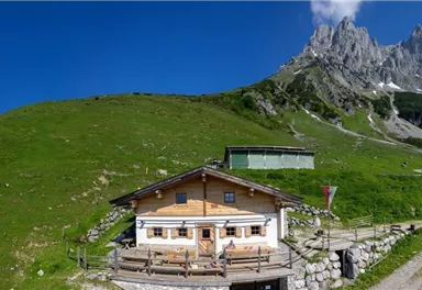 An alpine hut surrounded by green meadows and majestic mountains. The sky is clear and sunny, perfect for an outing in nature.