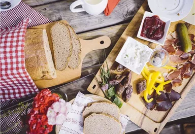 A rustic tray with fresh bread, cold cuts, and side dishes. Next to a cup of coffee, there’s a flowering branch and a decorative fabric.