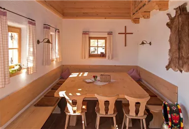 A cozy dining area with a large wooden table and benches. The walls are made of wood, and there are windows with curtains as well as a cross decoration.