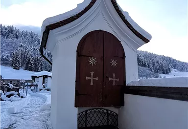 A snow-covered landscape with a small white building and a wooden door. In the background, there are snow-covered trees and a clear sky.