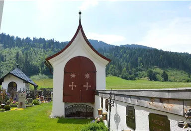 Eine kleine Kapelle mit einem roten Tor steht inmitten einer grünen Landschaft. Im Hintergrund sind sanfte Hügel und Bäume zu sehen.
