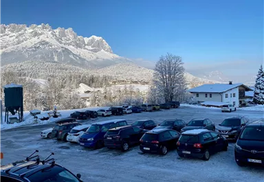 Ein Parkplatz mit mehreren Autos und einer verschneiten Landschaft im Hintergrund. Im Hintergrund sind Berge und ein klarer blauer Himmel zu sehen.