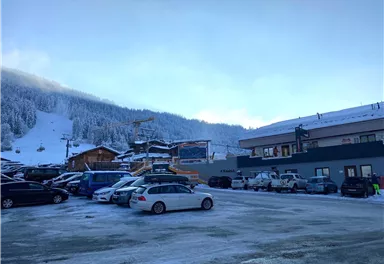 Ein Parkplatz in den Bergen, umgeben von schneebedeckten Hügeln. Im Hintergrund sind Skilifte und Gebäude zu sehen.