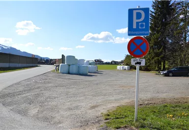 Ein Parkplatz mit einem Parkverbotsschild und runden Ballen im Hintergrund. Der Himmel ist blau mit einigen Wolken.