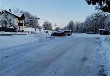 A snowy street with houses and trees in the background. The sky is clear and blue.