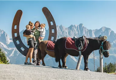 A family stands happily in front of a horseshoe gate. In the background, mountains can be seen, and next to them stands a small pony.