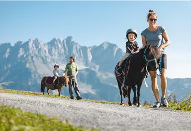 A woman is leading a pony with a small child on its back. In the background, there are mountains and a second family visible.