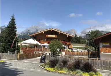 A traditional wooden house with a beautiful garden and mountains in the background. The sky is clear and sunny.