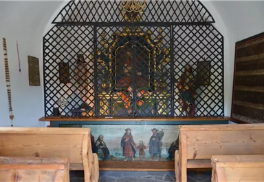 Eine kleine Kapelle mit Holzbankreihen und einem kunstvoll verzierten Altar. Die Wände sind schlicht, und die Fenster sind mit einem Gitter versehen.